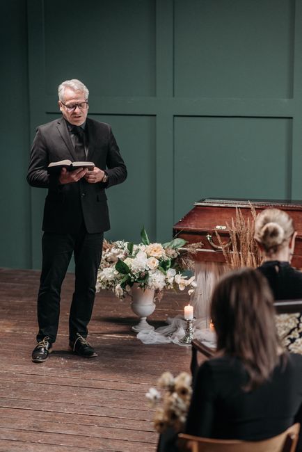 A man dressed in a black suit and glasses is reading from a book during a funeral service held in a room with dark green panelled walls. In front of him, there is a large floral arrangement composed o