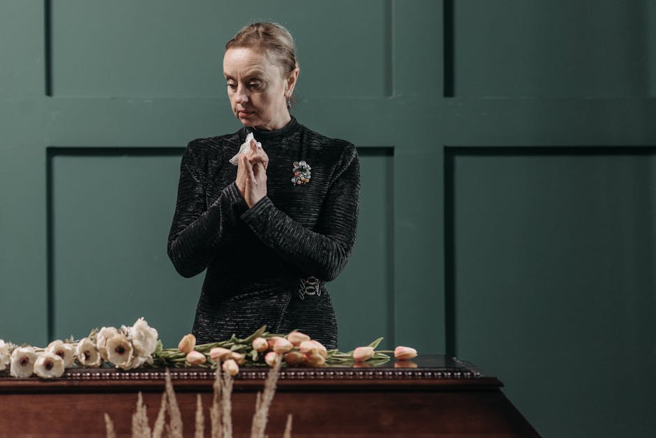 A woman dressed in a black textured top with a decorative brooch stands solemnly behind a dark wooden table, her hands clasped together in prayer or reflection. In front of her, arranged on the table,