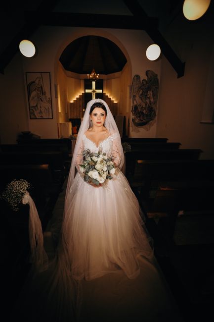 A bride standing inside a church, holding a floral bouquet featuring white roses, eucalyptus, and other mixed greenery. She is wearing a traditional white wedding gown with lace detailing and a long t